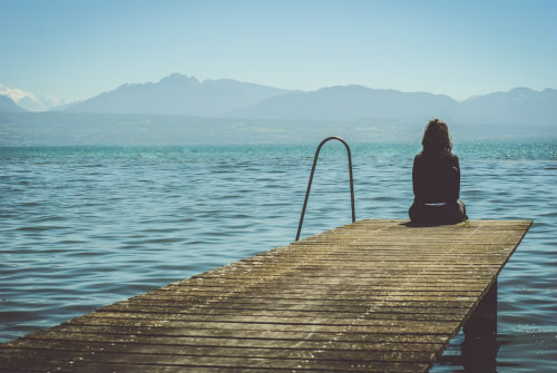 Girl sitting solo on a pier, looking out over the sea to mountains beyond, illustrating ‘how to love being alone’.