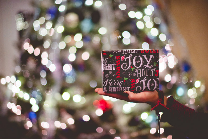 Gift paper with 'joy' in large letter, wrapping a gift in front of a blurred Christmas tree.