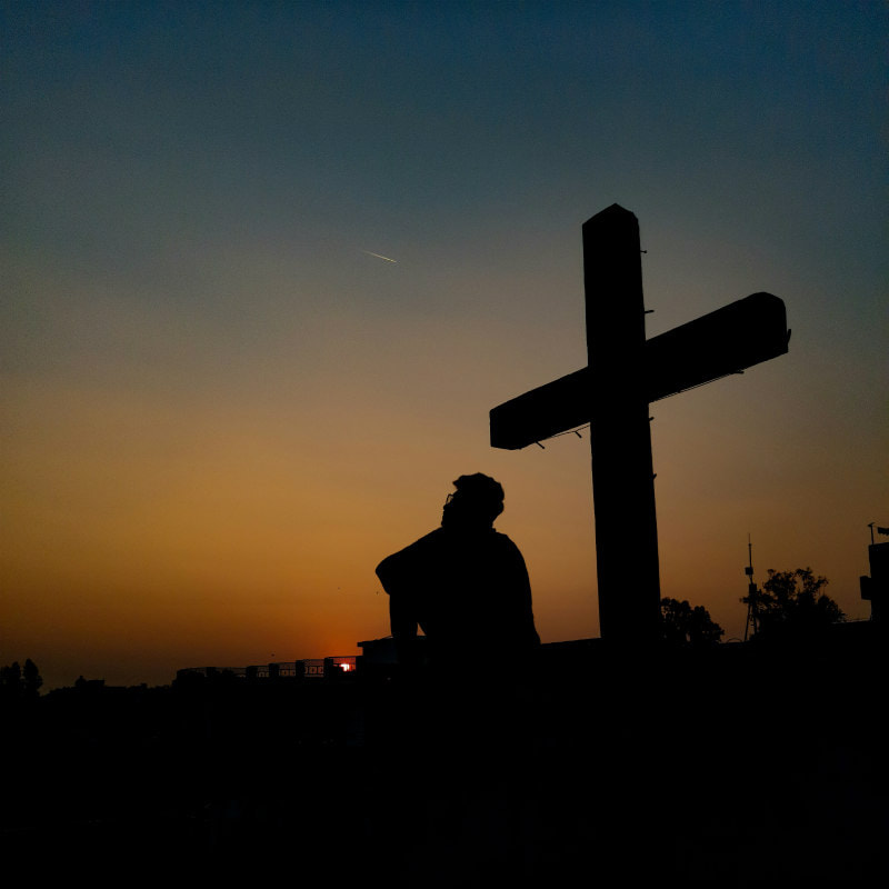 Person sitting gazing in the sunset, at the foot of a large wooden cross.
