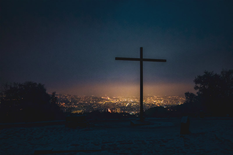 Cross on headland overlooking illuminated city, illustrating theme, 'Mighty is the power of the cross'.