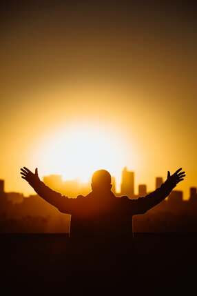 Man with hands outstretched, looking out over a sunset cityscape, illustrating the topic of ‘the Holy Spirit and Prayer’.