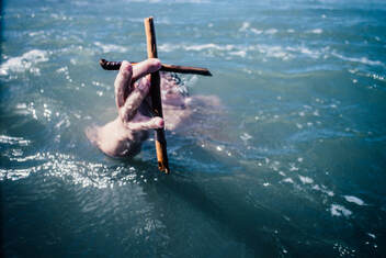 Hands holding a small cross above the surface of some water, illustrating baptism by immersion.