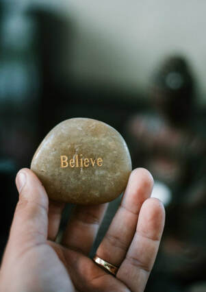 Man's hand holding pebble with 'believe' etched on it.