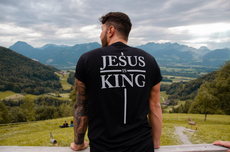 Man in black tee-shirt emblazoned with 'Jesus is King', looking out over a beautiful mountainscape.
